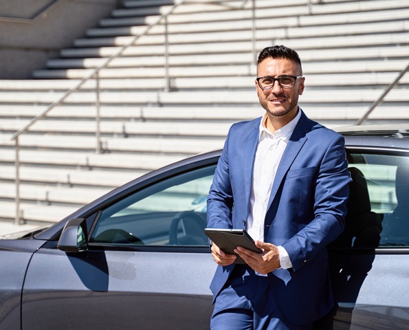 Smiling executive leaning on car, holding tablet and looking at camera Smiling executive leaning on car, holding tablet and looking at camera