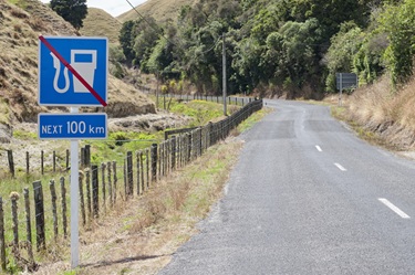 A rural New Zealand road winding through dry hills with a roadside sign indicating no fuel available for the next 100 kilometres.