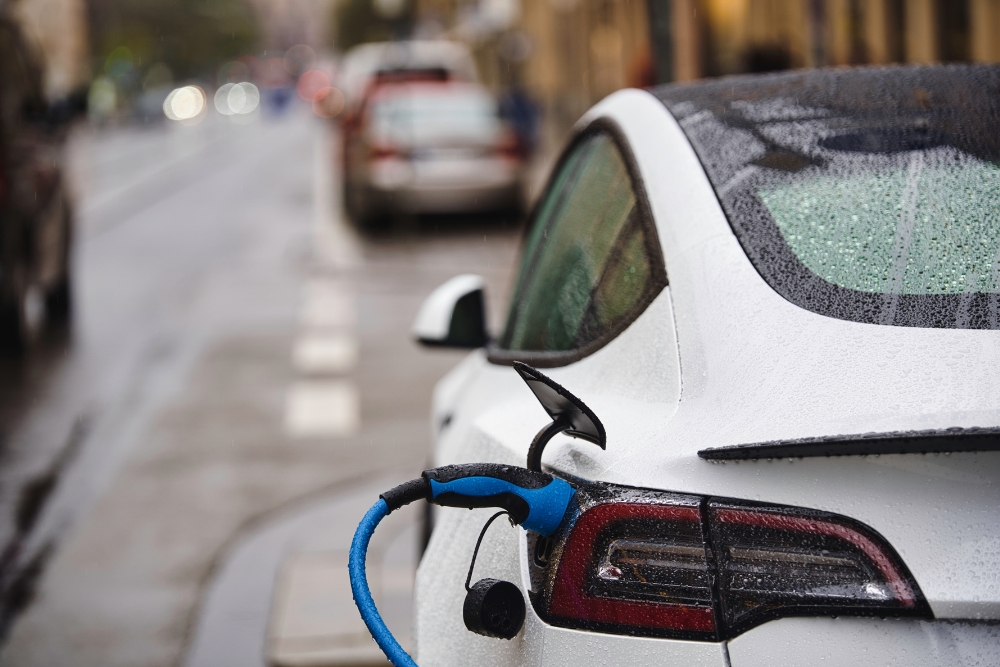 Are electric cars expensive: The rear of a wet white electric car parked on a rainy city street, with a blue charging cable connected to its charge port.