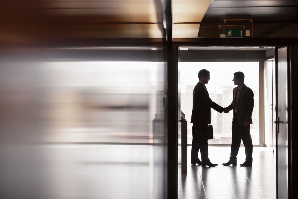 Automotive sector employee benefits Two business colleagues in suits shake hands in a backlit office corridor, illustrating employee benefits in the automotive sector.