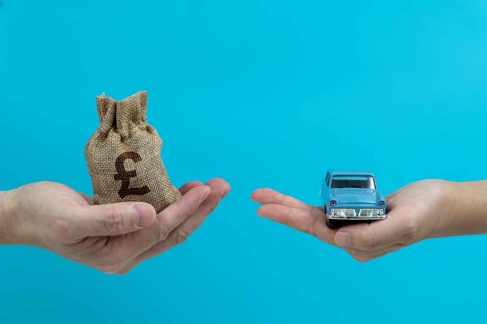 A hand holds a bag marked with a £ sign while another holds a small, blue toy car