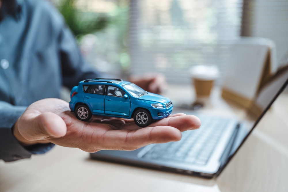 digital fleet administration tools: Hand holding a small blue model car in front of a laptop, symbolising digital tools and systems used for fleet management.