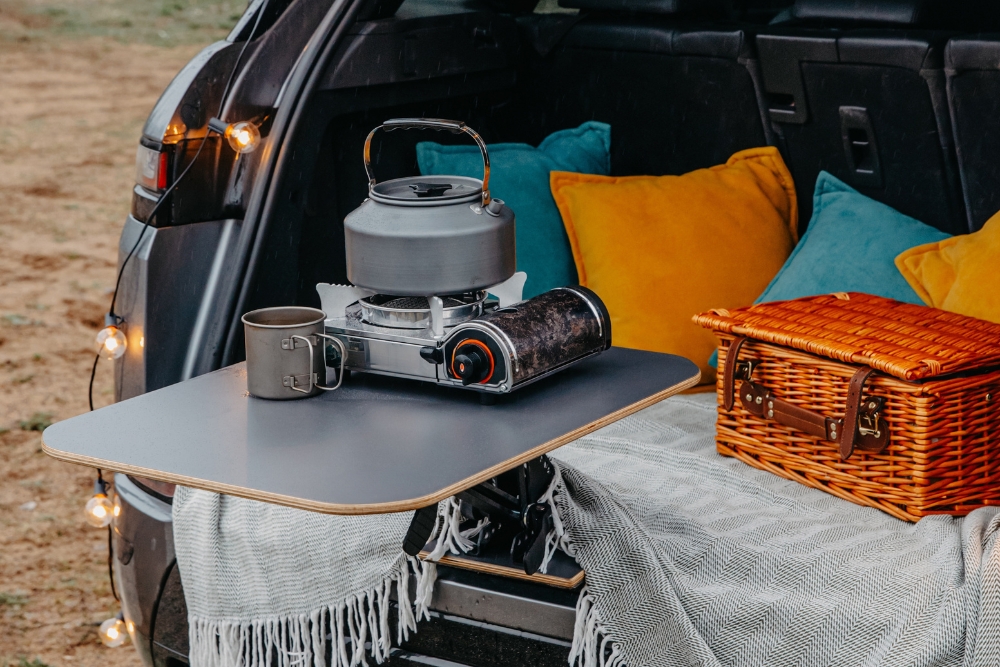 Boot of an SUV set up for camping with a picnic basket, cushions, and a portable stove on a bumper-mounted table.