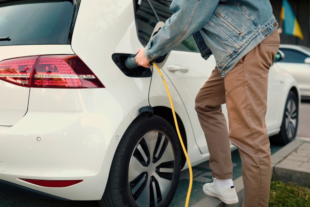 EV Prices UK: A person in a denim jacket and beige trousers plugging a yellow charging cable into the charge port of a white electric car in a car park.