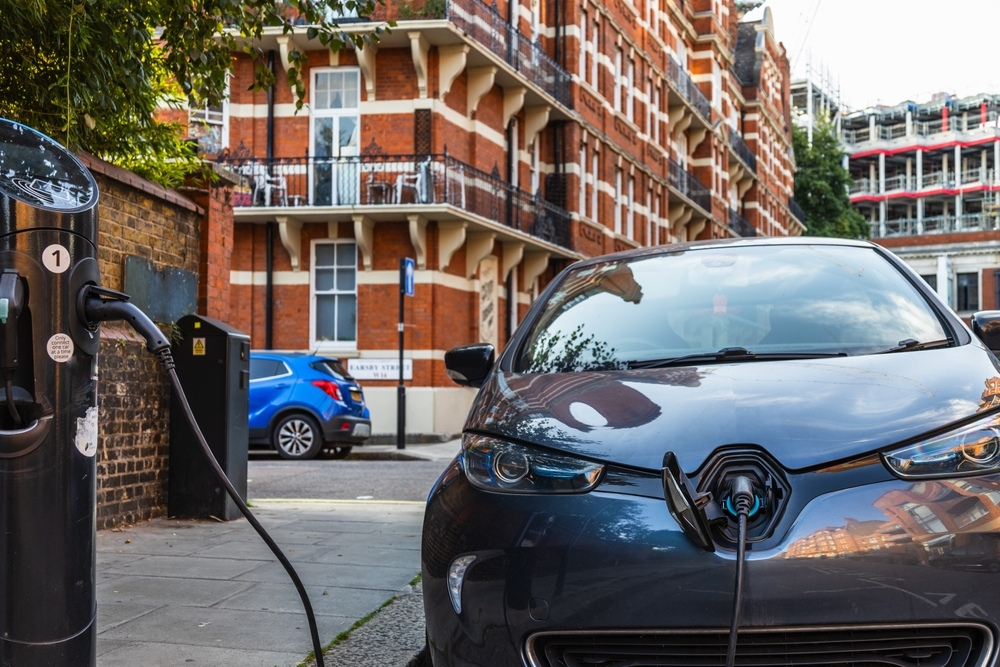 EV urban mobility Electric car plugged into a residential street charging station in front of red-brick housing in an urban area.