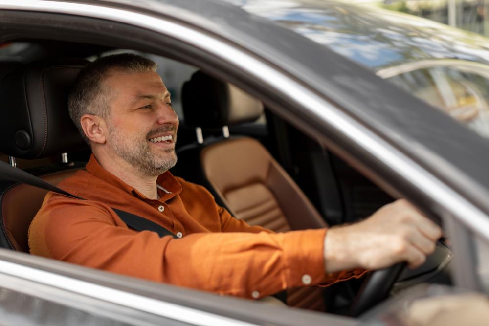 Hot weather commuting tips A smiling man driving a car with the window down, wearing an orange shirt.